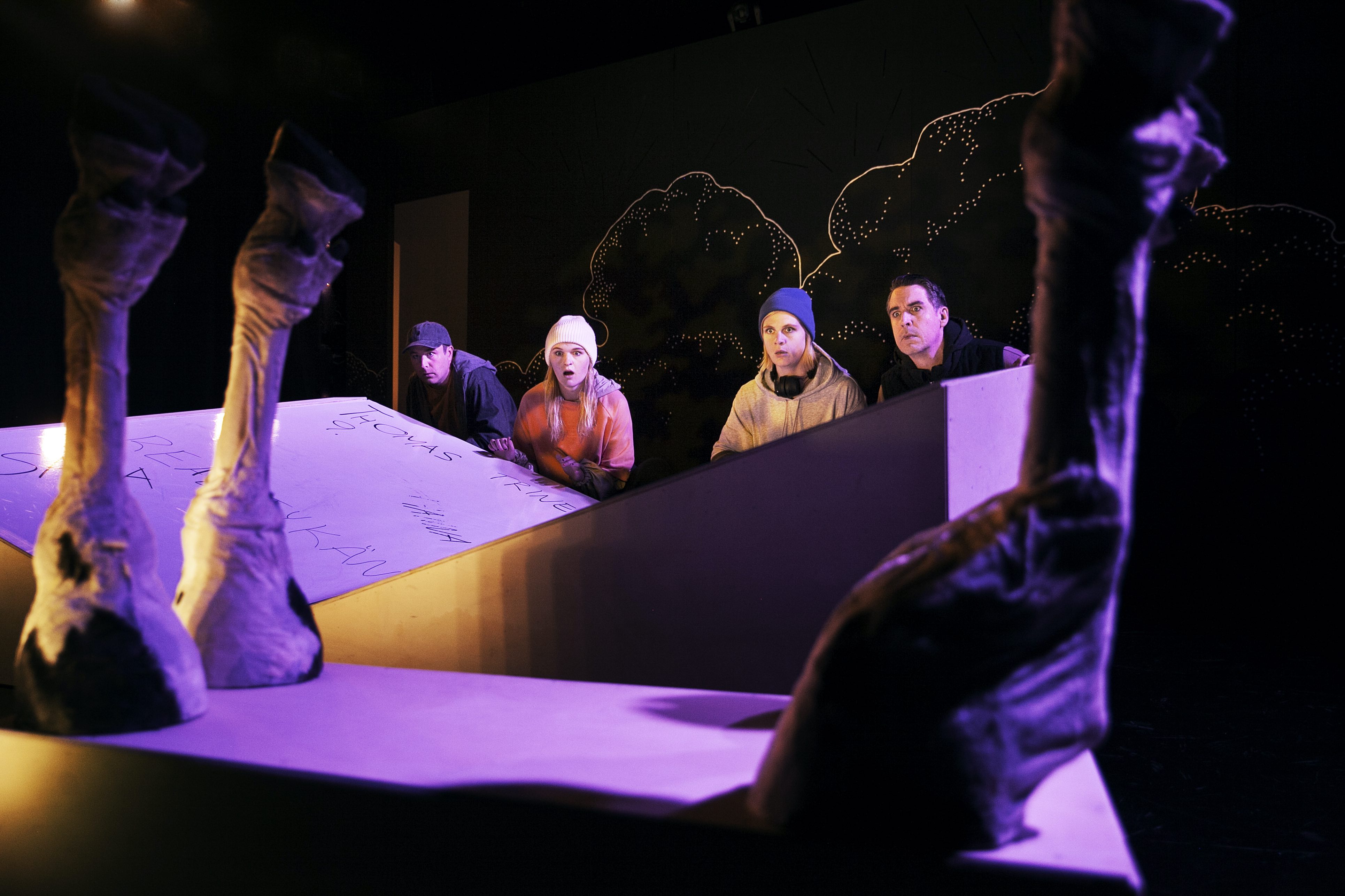 Foto på Thomas (Gustav Bloom), Trine (Cajsa Svensson), Anna (Josefin Larsson) och Henrik (Björn Löfgren) som tittar fram bakom scenografin och ser på fyra ko-ben som sticker upp i förgrunden.